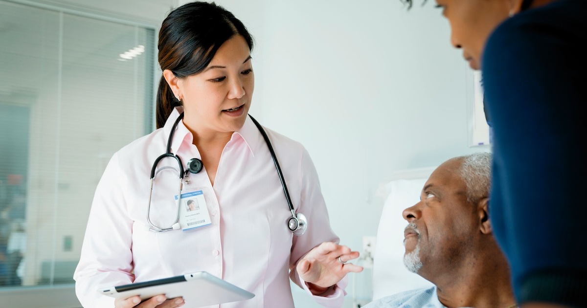 
  Doctor holding a tablet and standing by a patient and another individual 
