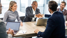 Two people sitting across from each other at a table and shaking hands while other watch