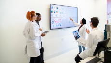 Professional team of medical scientists in a laboratory gathered around a wall monitor discussing charts and data