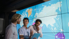 Three people standing in front of a board showing the world with lines connecting countries