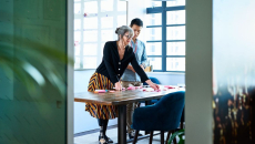 Two people standing in a room looking at something on a desk while standing above it