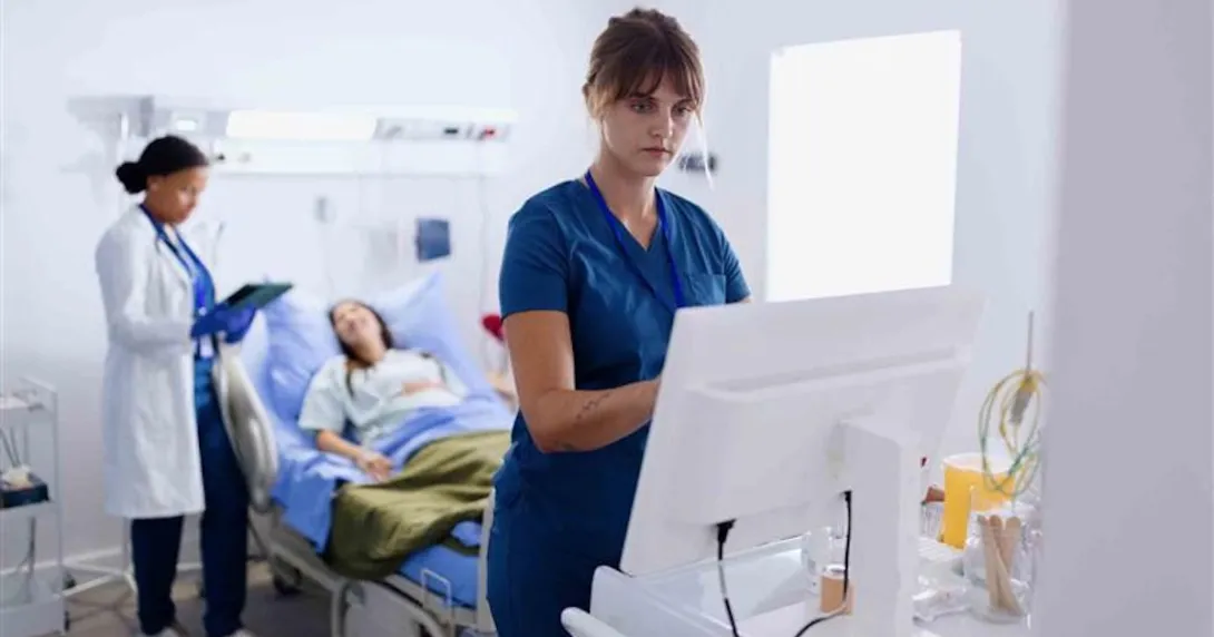 Nurse in a hospital setting looking at a computer while a doctor stands behind them and examines a patient in a hospital bed