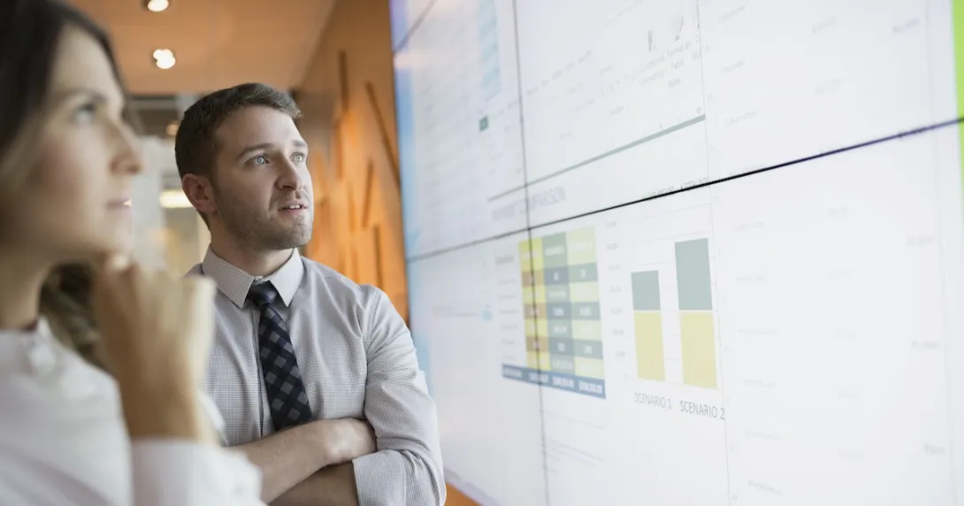 Two people standing up and looking at a large screen with graphs and numbers on it