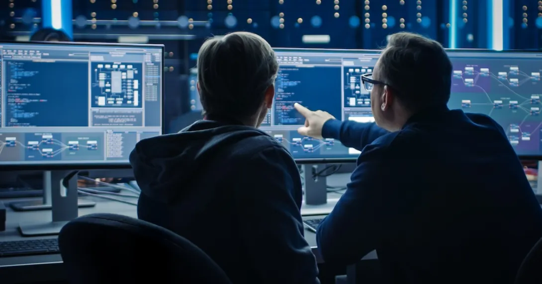 Two individuals sitting in front of two computer screens showing a plethora of data