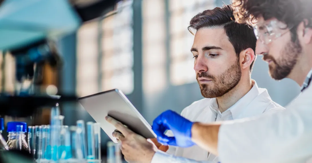 Two people in a laboratory looking at a tablet with beakers behind them