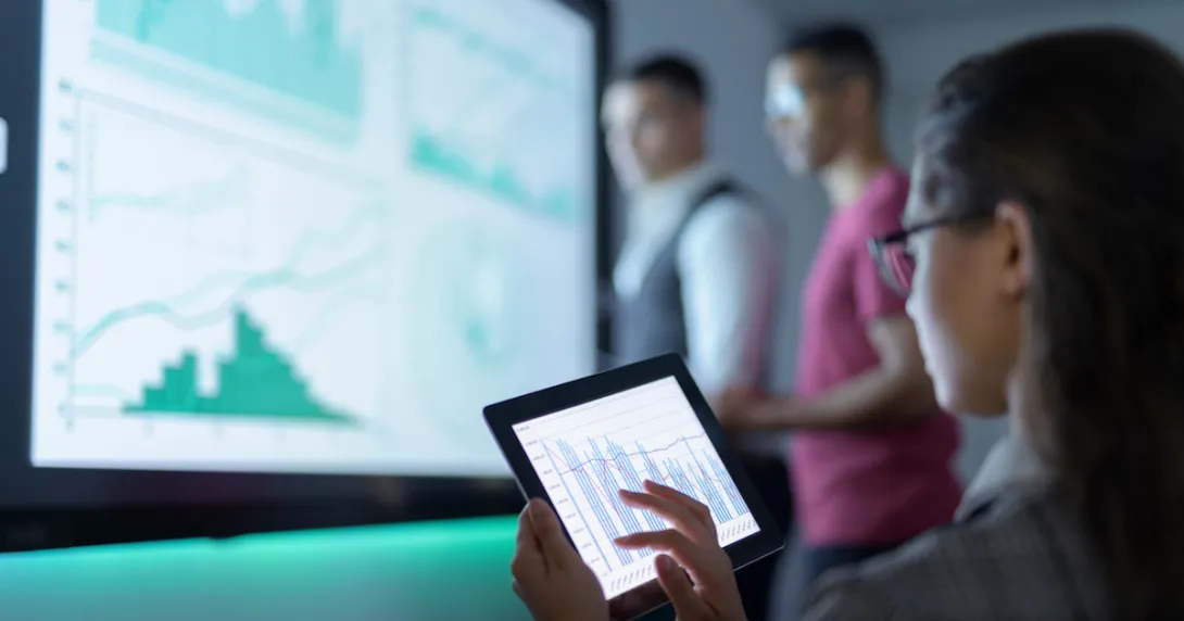 Two people standing in front of a screen looking at a graph and one person sitting down looking at a tablet