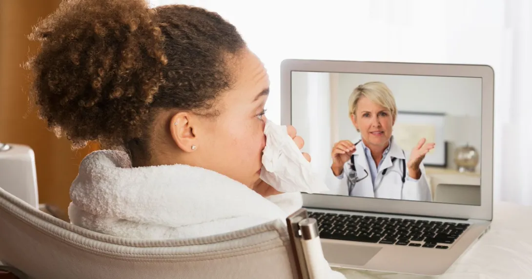 Person sitting in a chair while blowing their nose and talking to a healthcare provider on a computer