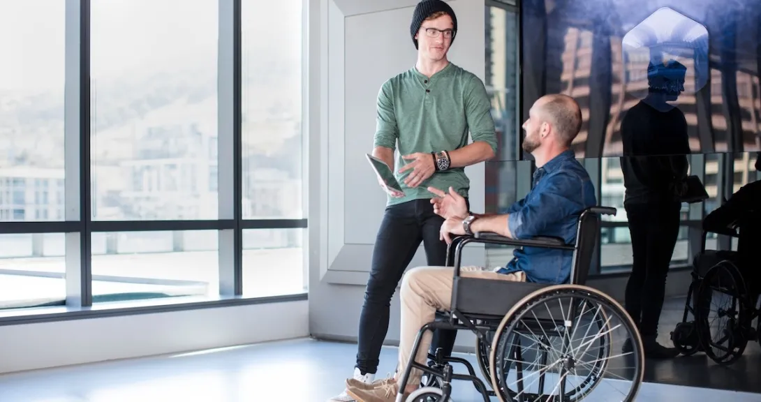Two people in a room next to a lot of windows with one in a wheelchair