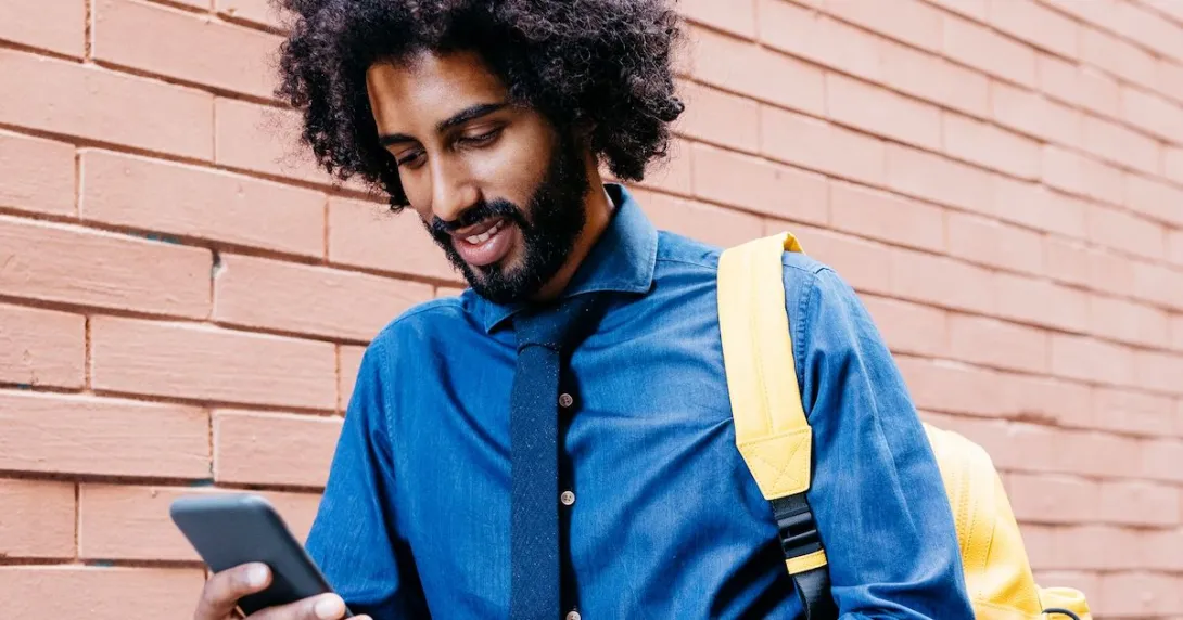 Person walking beside a brick wall wearing a blue shirt and yellow backpack while looking at a phone