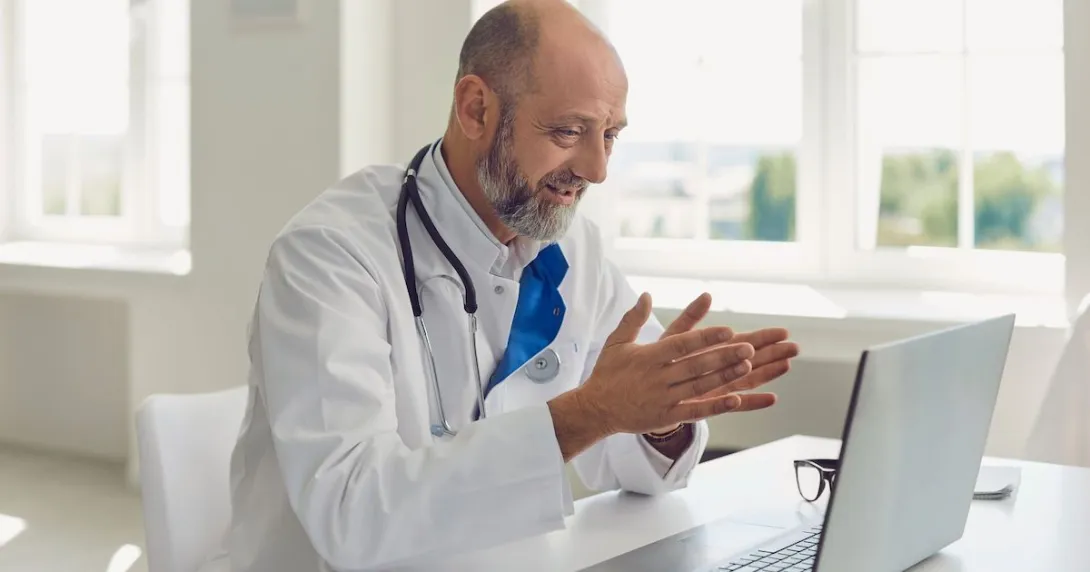 Healthcare provider wearing a lab coat and a stethoscope around their neck while sitting at a desk looking at a computer