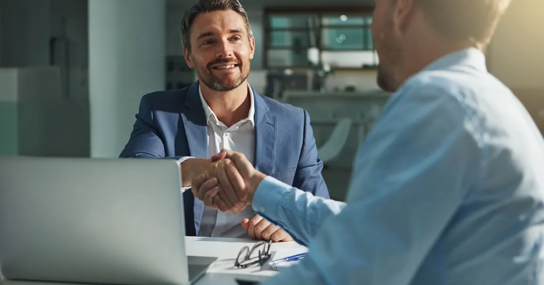 Two people sitting on opposite sides of a desk shaking hands