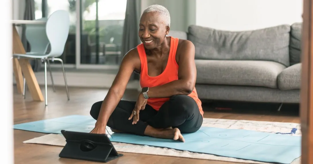 An older woman sitting on a yoga mat looking at exercises on her tablet