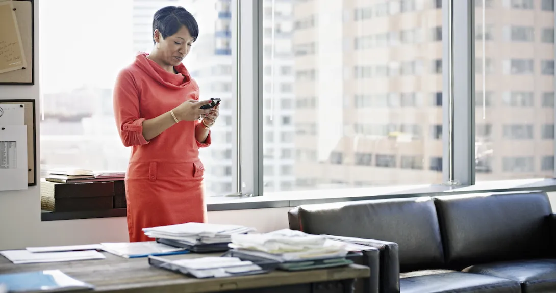 Businesswoman standing in office looking at smart phone smiling